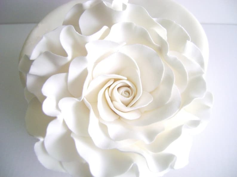 Puede incluir: Una gran rosa blanca de fondant con p&eacute;talos rizados sobre un pastel blanco.
