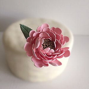 May include: A pink sugar flower with a gold center and green leaves. The flower is on top of a white cake.