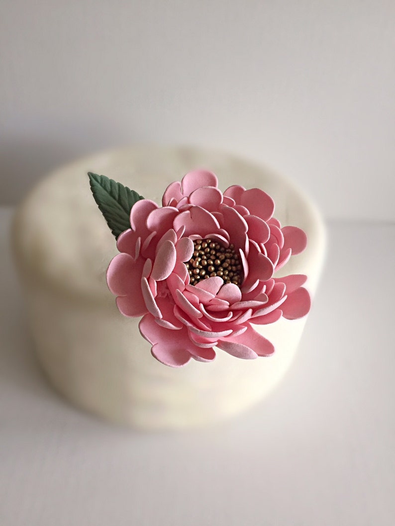 May include: A pink sugar flower with a gold center and green leaves. The flower is on top of a white cake.