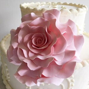 Op de afbeelding: Een witte bruidstaart met twee lagen, versierd met gepipete frosting en een grote, roze fondantroos.
