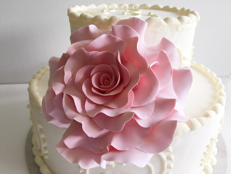 Op de afbeelding: Een witte bruidstaart met twee lagen, versierd met gepipete frosting en een grote, roze fondantroos.