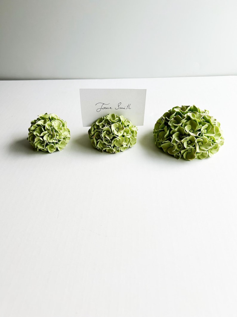 Puede incluir: Tres porta tarjetas de lugar con forma de flor de hortensia verde. El porta tarjetas del medio tiene una tarjeta blanca con el nombre "Jane Smith" escrito en ella.