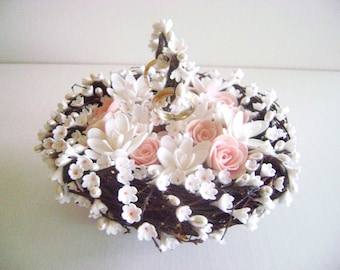 Op de afbeelding: Een wit en roze bloemen kussen voor ringen met twee gouden ringen erop. Het kussen is gemaakt van witte en roze bloemen en is omringd door kleine witte bloemen.