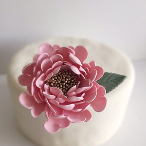 May include: A pink sugar flower with gold accents sits on top of a white cake.