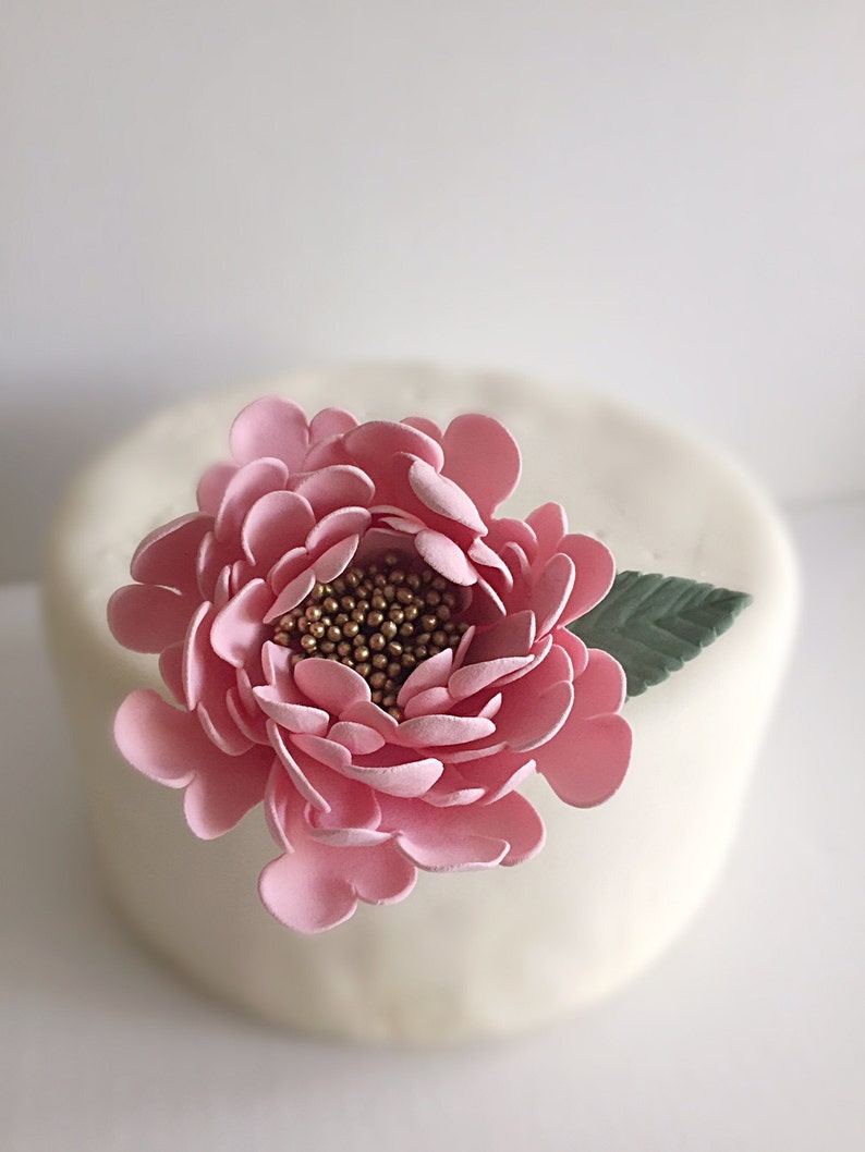 May include: A pink sugar flower with gold accents sits on top of a white cake.