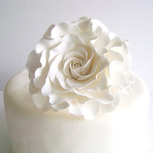 Puede incluir: Una rosa blanca de fondant sobre un pastel blanco. La rosa est&aacute; hecha de pasta de az&uacute;car y tiene una textura delicada y rizada.
