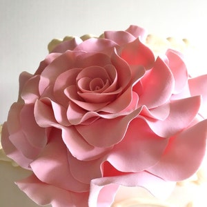 Op de afbeelding: Een grote, roze fondantroos zit bovenop een witte taart. De roos is gemaakt van vele lagen bloemblaadjes, waardoor een volle en elegante uitstraling ontstaat.