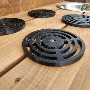 K&ouml;nnte beinhalten: Vier schwarze, runde Abflussabdeckungen aus Kunststoff mit konzentrischem Ringdesign, auf einer Holzoberfl&auml;che platziert.