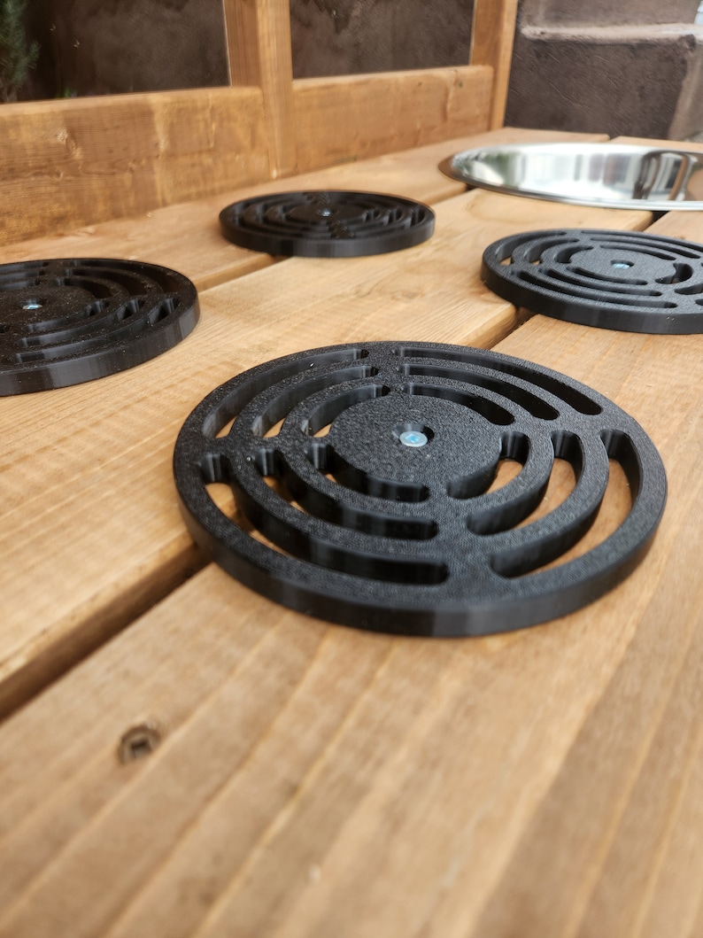 K&ouml;nnte beinhalten: Vier schwarze, runde Abflussabdeckungen aus Kunststoff mit konzentrischem Ringdesign, auf einer Holzoberfl&auml;che platziert.