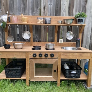 K&ouml;nnte beinhalten: Holzspeisek&uuml;che f&uuml;r den Au&szlig;enbereich mit Sp&uuml;le, Herd und Backofen. Die K&uuml;che hat Regale zum Verstauen und Haken zum Aufh&auml;ngen von Utensilien. Auf den unteren Regalen befinden sich schwarze Plastik-Aufbewahrungsbeh&auml;lter. Die K&uuml;che ist braun lackiert und hat einen rustikalen Look.