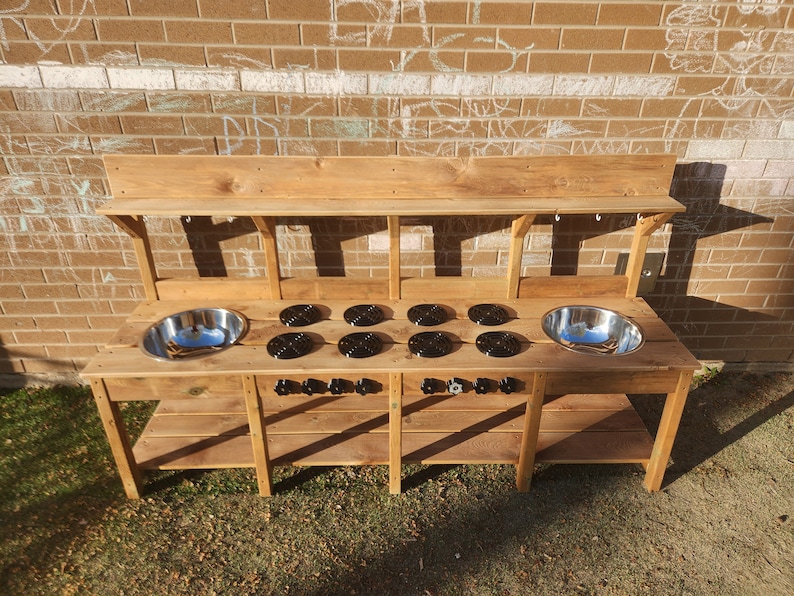 K&ouml;nnte beinhalten: Eine h&ouml;lzerne Spielk&uuml;che mit zwei Edelstahlsp&uuml;len, sechs schwarzen Herdplatten und zwei Regalen. Die K&uuml;che ist aus Naturholz gefertigt und hat einen rustikalen Look.