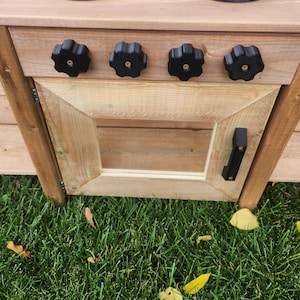 May include: A wooden play kitchen with black knobs and a door with a black handle. The kitchen is made of light-coloured wood and is set on a grassy surface.