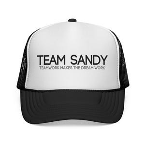 May include: A black and white trucker hat featuring the text "TEAM SANDY" in black. Underneath, the hat reads "TEAMWORK MAKES THE DREAM WORK". The hat has a black peak and a mesh back for breathability.