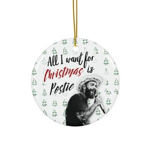 May include: A white ceramic Christmas ornament with a gold string for hanging. The ornament features a black and white image of a singer and the text "All I want for Christmas is Postie" in black and red.