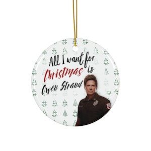 May include: A white ceramic Christmas bauble with a green and white tree pattern. The bauble features a photo of Owen Strand from the TV show "9-1-1: Lone Star" and the text "All I want for Christmas is Owen Strand".