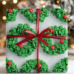 May include: A wrapped gift with a festive design. The wrapping paper features a pattern of green wreath-shaped treats with red candies on a white marble background. A red ribbon and bow adorn the gift, perfect for the holiday season.