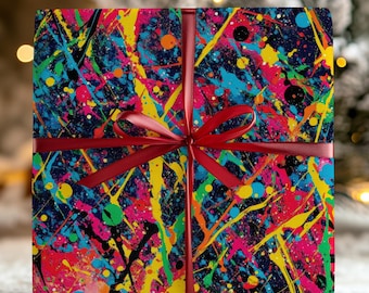 May include: A wrapped gift box featuring a vibrant, abstract design of colorful paint splatters on a dark background. The gift is tied with a red ribbon and bow. The background includes a snowy surface and a blurred Christmas tree.