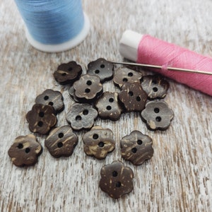 May include: A collection of flower-shaped, two-hole buttons made from dark brown coconut shell. A spool of light blue thread and a spool of pink thread with a needle are in the background, suggesting sewing or crafting.
