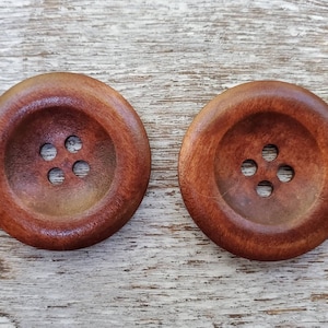 May include: Two large, round wooden buttons with a rich, reddish-brown hue. Each button features four evenly spaced holes for sewing. The buttons are set against a light, weathered wooden surface, highlighting their natural grain and texture.