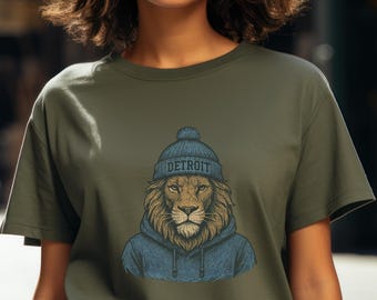 May include: Olive green t-shirt featuring a lion wearing a blue beanie with the word "DETROIT" and a blue hoodie. The lion has a detailed mane and is centered on the shirt. The shirt has short sleeves.