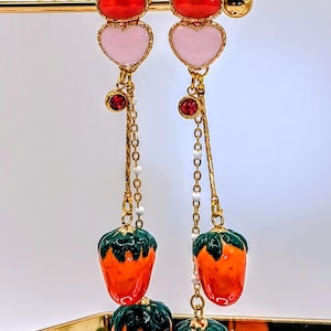 Peut inclure: Une paire de boucles d'oreilles pendantes avec un design de cœur superposé. Le cœur supérieur est rouge, le cœur du milieu est rose et le charme du bas est une fraise orange à feuilles vertes.