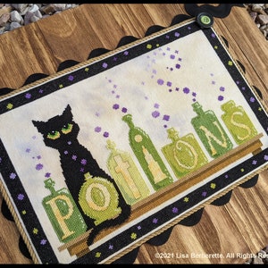 May include: Cross-stitch artwork featuring a black cat with green eyes and the word "POTIONS" above a shelf of potion bottles. The artwork is framed with a black scalloped border and green buttons, set on a wooden background.