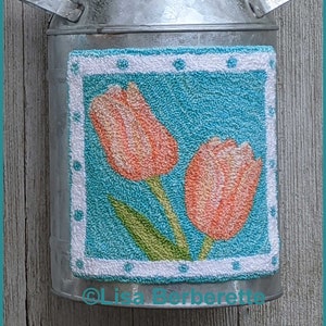 May include: A galvanized metal milk can-shaped vase with a turquoise and white floral design. The design features two orange tulips with green leaves. The vase has a flared top and two handles.