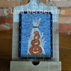 Może przedstawiać: Oprawione dzieło sztuki tekstylnej przedstawiające stos trzech rzeźbionych dyni z uśmiechniętymi twarzami, na tle niebieskiego nieba. Grafika jest prezentowana na drewnianym stojaku w stylu deski do krojenia. Tekst na obrazku brzmi "Sweet Stack PDF Pattern".