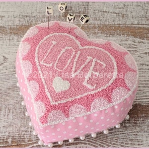 May include: A pink heart-shaped pincushion with the word "LOVE" embroidered in white. The pincushion has a textured surface and is decorated with white polka dots and a border of pearls. Small lettered pins spell out "LOVE".