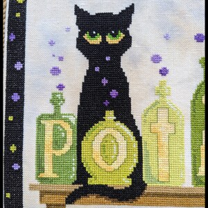 May include: Cross-stitch artwork featuring a black cat with green eyes, surrounded by green potion bottles. The word "POTION" is spelled out on the bottles. Purple star-shaped accents and a black border complete the design.
