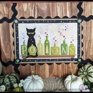 May include: Cross-stitch artwork featuring a black cat with green eyes sitting in front of potion bottles. The word "POTIONS" is displayed in green letters. The artwork is framed with a wooden border and black accents, perfect for Halloween decor.
