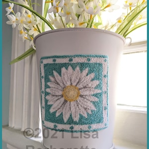 Op de afbeelding: Witte metalen emmer met een madeliefjesontwerp. De emmer heeft een vierkante, turquoise en witte madeliefjesapplicatie. Het madeliefje heeft een geel hart en witte bloemblaadjes. De emmer is gevuld met witte bloemen.
