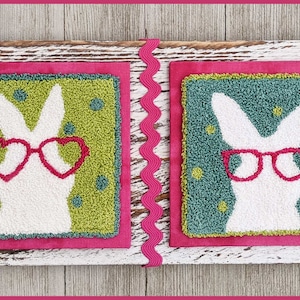 Puede incluir: Un letrero decorativo de madera con dos paneles cuadrados texturizados. Cada panel presenta una silueta de conejo blanco con gafas rosas en forma de corazón. Los paneles están sobre un fondo rosa con un borde verde y azul, y un ribete de cinta rosa.