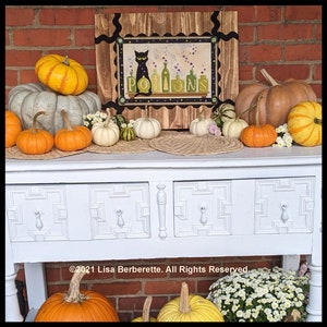 May include: A Halloween-themed display featuring various pumpkins in shades of orange, yellow, and white, arranged on a white table. A framed artwork with a black cat and the word "POTIONS" is the centerpiece. Decorative gourds and flowers complete the festive arrangement.