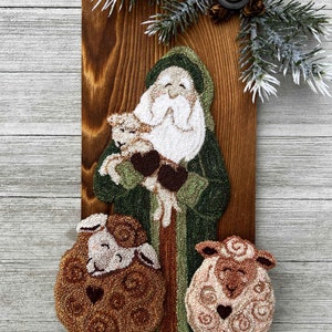 May include: A wooden wall hanging features a Santa Claus figure holding a lamb, with two sheep below. The figures are made of textured fabric in shades of green, brown, and white. A sprig of pine and a button adorn the top.