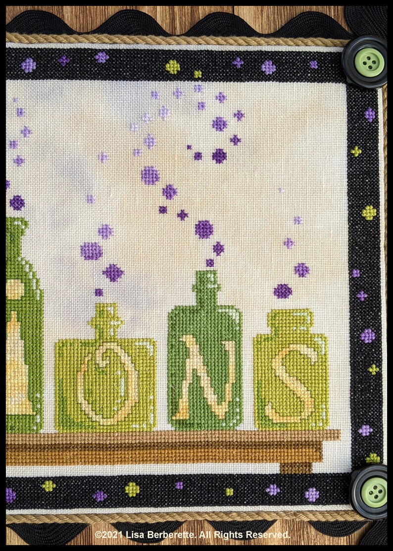 May include: Cross-stitch artwork featuring green and yellow potion bottles with the letters "IONS". Purple bubbles and stars float upwards. The border is black with green buttons and a rope trim. The artwork is framed.