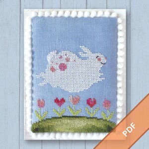 May include: Cross-stitch design featuring a white bunny with pink spots leaping across a light blue background. Below, a green grassy area displays pink tulips and hearts. The design is framed with a white border and pom-poms.