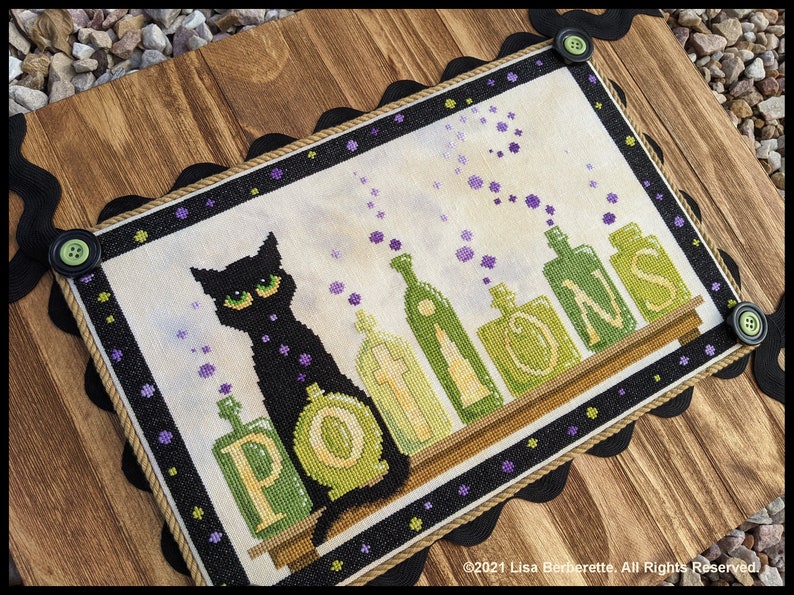 May include: Cross-stitch artwork featuring a black cat with green eyes and the word "POTIONS" above a shelf of potion bottles. The artwork is framed with a black scalloped border and green buttons, set on a wooden background.