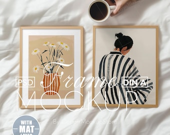 May include: Two framed illustrations on a white bedspread. The illustration on the left is a vase of white flowers with a light brown background. The illustration on the right is a black and white striped illustration of a person with their back turned. The text "Frame Mockup" is visible in the image.