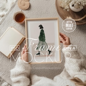 Puede incluir: Una maqueta de marco de madera con una capa de passe-partout, que presenta a una mujer con un vestido verde y blanco caminando. El marco está sostenido por una persona que lleva un suéter blanco. El texto "Frame Mockup" está superpuesto en la imagen.