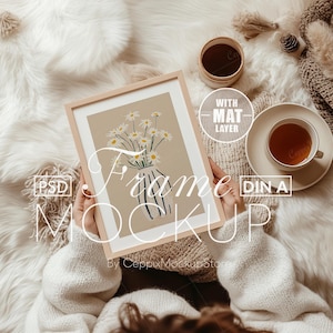 May include: A white wooden frame mockup with a mat layer, featuring a floral illustration of white daisies in a blue and white striped vase. The frame is held by a person wearing a white sweater.