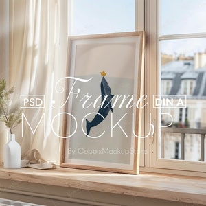 Peut inclure: Une impression encadrée d'une baleine bleue avec une couronne jaune sautant hors de l'eau. Le cadre est en bois et a une finition brun clair. L'impression est accrochée à un mur dans une pièce avec une fenêtre. La fenêtre est ouverte et il y a une vue sur une ville à l'extérieur. Le texte "Frame Mockup" est écrit sur l'image.