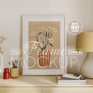 Puede incluir: Una maqueta de marco blanco con una capa de paspartú, que presenta una impresión de margaritas blancas en un jarrón naranja a rayas. El marco está sobre una mesa de madera clara con una lámpara, un jarrón con flores secas y un porta lápices con lápices.