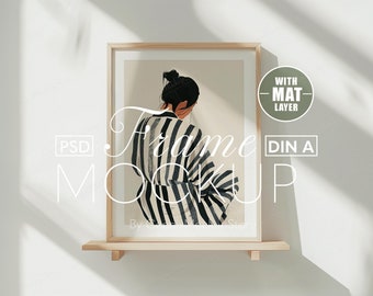 May include: A framed art print mockup featuring a woman in a black and white striped garment. The artwork is displayed in a light wood frame with a white mat, resting on a small wooden shelf. The text "Frame Mockup" is overlaid on the image.