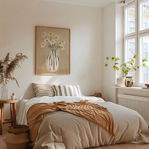 May include: A cozy bedroom scene with a bed covered in neutral-toned bedding and a brown throw blanket. A framed artwork of daisies in a vase hangs above the bed. A wooden stool, baskets, and a large window complete the inviting space.