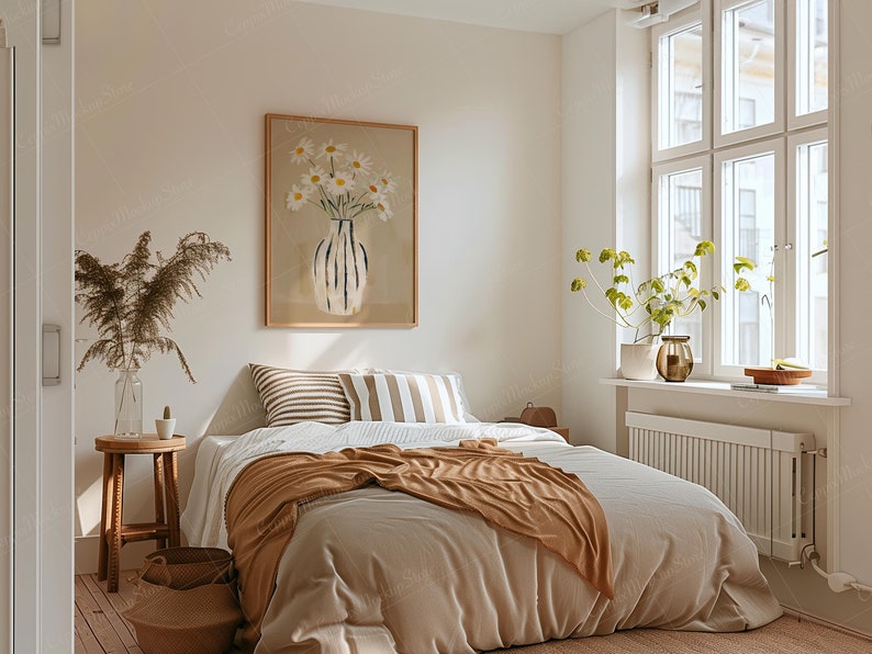 May include: A cozy bedroom scene with a bed covered in neutral-toned bedding and a brown throw blanket. A framed artwork of daisies in a vase hangs above the bed. A wooden stool, baskets, and a large window complete the inviting space.