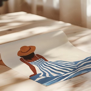 May include: Rolled art print featuring a woman in a blue and white striped dress and a brown hat. The illustration depicts a person relaxing in a sunny setting. The artwork is displayed on a wooden surface.