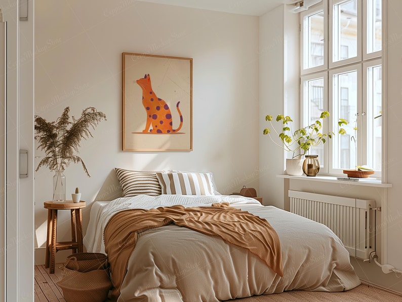 May include: A bedroom scene featuring a bed with neutral-toned bedding and a brown throw blanket. A framed artwork of an orange cat with purple spots hangs above the bed. The room is decorated with a wooden stool, a vase of dried plants, and a window with natural light.
