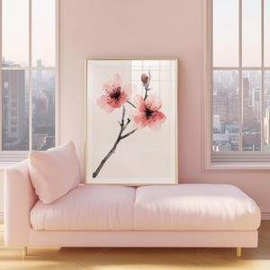 May include: A framed floral art print featuring pink cherry blossoms with black stems and a gold frame. The artwork is displayed above a pink chaise lounge with a matching pillow, set against a pink wall and large windows.