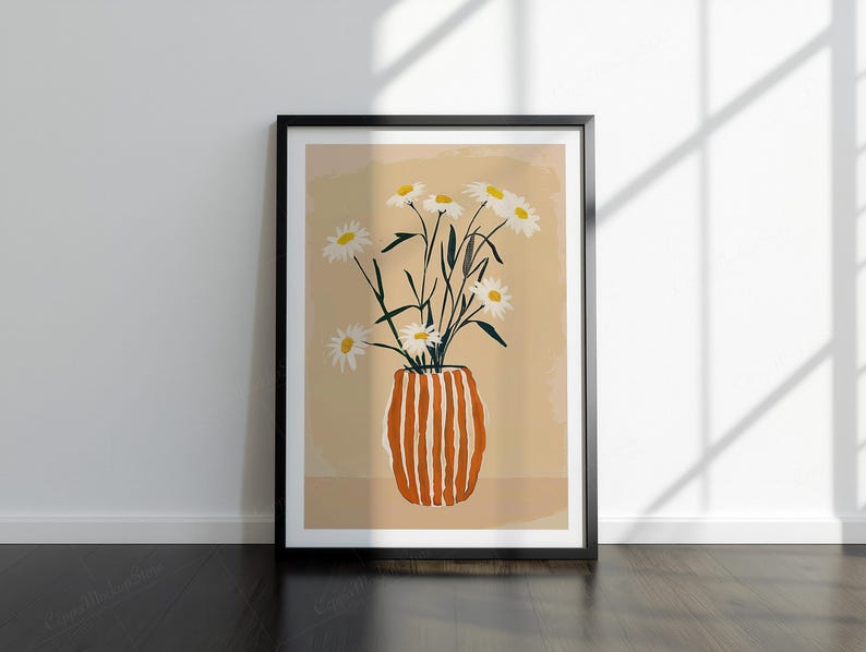 May include: Framed art print showing a vase of white daisies with green stems. The vase is orange with vertical white stripes. The background is a light tan colour. The frame is black and the artwork is leaning against a white wall.